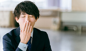 生活習慣のイメージ｜デスクに座ったスーツ姿の男性が眠そうに口元を押さえている様子